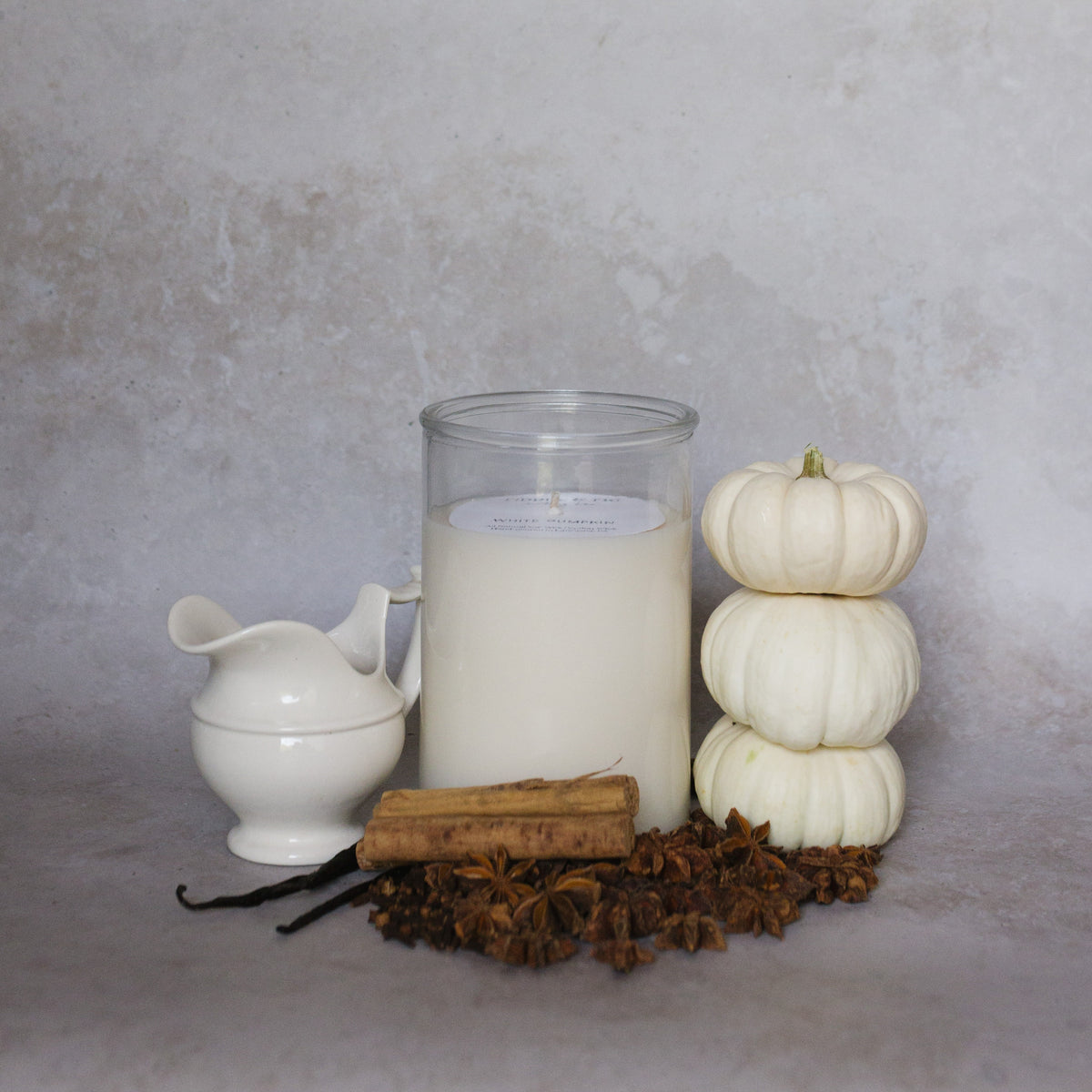A large clear glass jar candle filled with white wax stands on a neutral surface, styled with a small white ceramic pitcher, cinnamon sticks, star anise, vanilla beans, and three stacked small white pumpkins against a soft textured gray background.